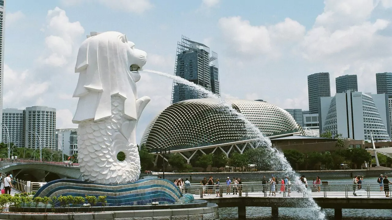 The Merlion fountain in Singapore, featuring a lion's head and fish body, spouts water against a city skyline backdrop.