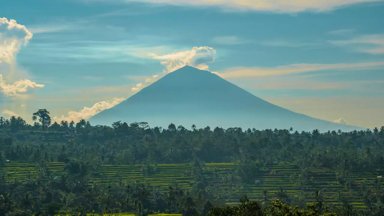 A large mountain rises against a backdrop of fluffy clouds in a clear blue sky.