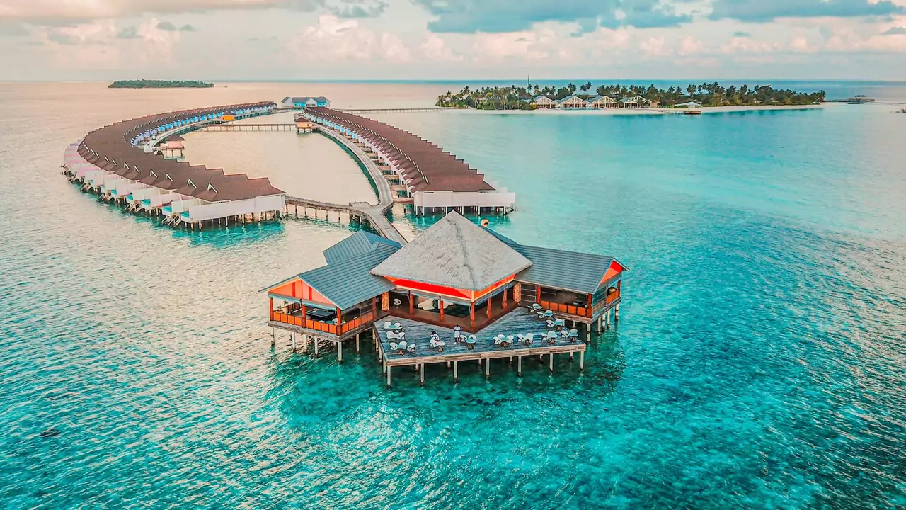 Aerial view of a tropical resort surrounded by clear blue ocean waters, showcasing palm trees and sandy beaches.