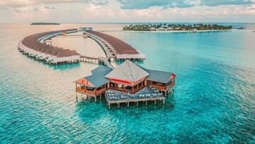 Aerial view of a tropical resort surrounded by clear blue ocean waters, showcasing palm trees and sandy beaches.