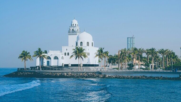 A white mosque by the ocean, surrounded by sand and water, creating a serene coastal scene.