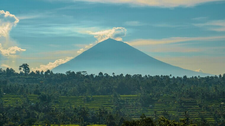 A large mountain rises against a backdrop of fluffy clouds in a clear blue sky.