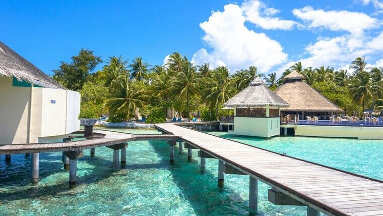 A wooden pier extends to a tropical resort featuring thatched huts surrounded by water and greenery.